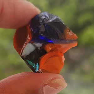 Puede incluir: Un trozo de vidrio colorido de forma irregular, con una mezcla de tonos naranja, azul, blanco y verde azulado. El vidrio se sostiene entre dos dedos, mostrando su superficie texturizada única. El fondo está borroso, sugiriendo un entorno al aire libre.