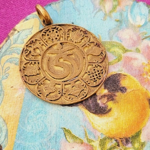 May include: A gold-colored pendant with an intricate design featuring a central symbol and surrounding patterns. The pendant rests on a decorative, round coaster with a floral and bird design in shades of blue, pink, and yellow. The background is a vibrant magenta fabric.