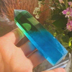 Puede incluir: Un largo cristal de resina transparente con la parte superior verde y la inferior azul. El cristal tiene forma de punta y se sostiene en una mano.