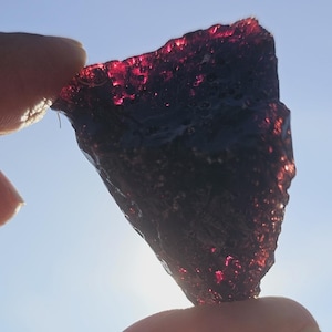 May include: A dark red, irregularly shaped gemstone held between fingers against a blue sky. The stone has a rough, textured surface with sparkling highlights, suggesting a crystalline structure. The overall appearance is translucent.