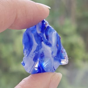 Könnte beinhalten: Ein Stück blaues und weißes Glas, das zwischen zwei Fingern gehalten wird. Das Glas hat eine raue, unregelmäßige Form und erscheint durchscheinend. Die Farben sind ineinander gewirbelt, wodurch ein Marmoreffekt entsteht. Der Hintergrund ist unscharf.