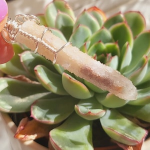 Puede incluir: Una punta de cristal blanco envuelta en alambre plateado, descansando sobre una planta suculenta verde en una maceta blanca.