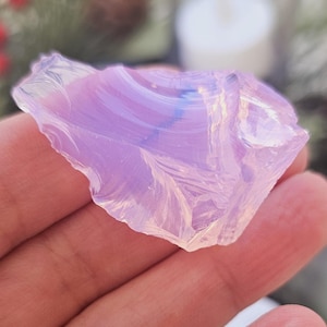 Könnte beinhalten: Ein durchscheinender, unregelmäßig geformter, hellvioletter Kristall, der in einer Hand gehalten wird. Der Kristall hat ein geschichtetes Aussehen mit Andeutungen von Blau und Weiß. Der Hintergrund ist verschwommen, mit Andeutungen von Grün und festlichen Dekorationen.