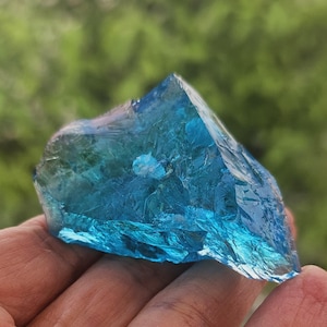 May include: A vibrant, translucent blue crystal fragment with a rough, textured surface. The crystal is held in a hand, showcasing its natural, irregular shape and colour variations. The background is blurred.