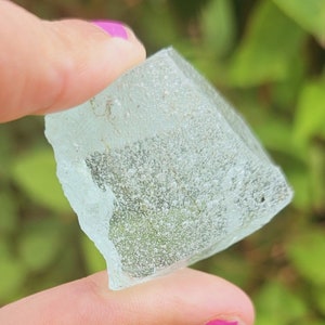 Op de afbeelding: Een helder, lichtblauw, gefacetteerd kristal met een ruwe, ongelijkmatige textuur. Het kristal is kubusvormig en wordt vastgehouden door een hand.