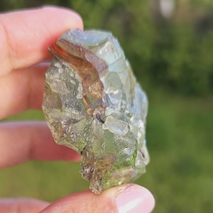 Könnte beinhalten: Ein durchscheinender, unregelmäßig geformter Kristall mit hellgrüner Tönung und braunen Einschlüssen. Der Kristall wird zwischen Fingern vor einem verschwommenen grünen Hintergrund gehalten, was seine natürliche, rohe Form zeigt. Die Oberfläche des Kristalls reflektiert Licht.