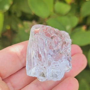 May include: A clear, faceted crystal with a rough, irregular shape. The crystal is held in a hand with pink nail polish.