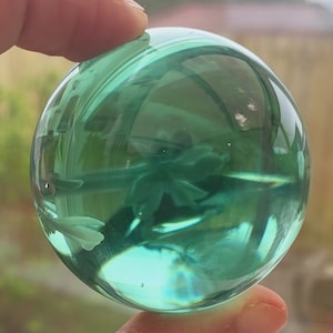 Puede incluir: Una esfera de vidrio transparente de color verde con un patrón de remolino en su interior. La esfera está siendo sostenida por un dedo.