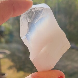 May include: A clear, translucent, white crystal with a rough, irregular shape. The crystal is being held by a hand.