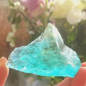 May include: A large piece of sea glass in a light turquoise color. The glass has a rough, textured surface and is translucent.