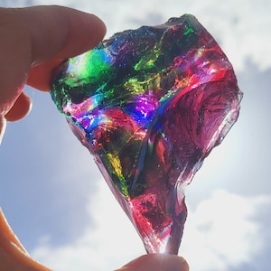 Puede incluir: Un gran fragmento de vidrio iridiscente, morado, verde y azul, con una forma irregular y rugosa. El fragmento se sostiene en una mano contra un cielo nublado.