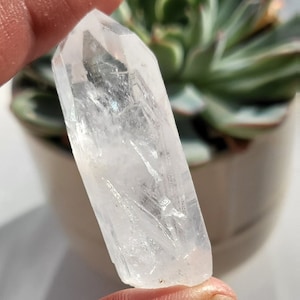 Puede incluir: Una punta de cristal de cuarzo transparente con una superficie lisa y pulida. El cristal se sostiene en una mano, y el fondo está desenfocado.