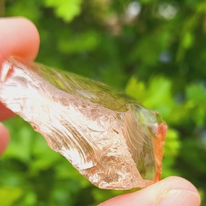 Puede incluir: Un cristal transparente, translúcido y ligeramente rosado con una forma irregular y rugosa. El cristal está siendo sostenido por una mano contra un fondo verde borroso.