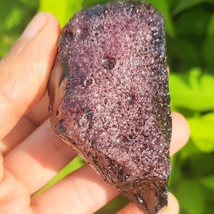 Puede incluir: Una pieza de obsidiana de color morado oscuro, de forma irregular y con una superficie texturizada. La obsidiana tiene muchas pequeñas burbujas de aire atrapadas en su interior.