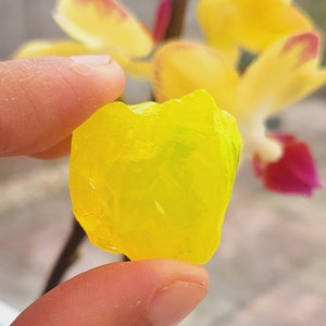 Puede incluir: Un cristal translúcido de forma irregular, de color amarillo brillante. El cristal está siendo sostenido por una mano.