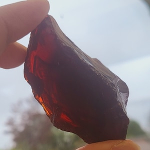 Puede incluir: Un trozo de vidrio rojo oscuro de forma irregular, probablemente obsidiana, sostenido en una mano.