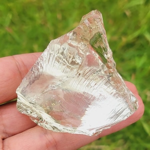 Puede incluir: Un trozo de vidrio transparente, translúcido, de forma irregular con un ligero tono verde. El vidrio tiene una superficie rugosa y texturizada y parece ser un trozo de vidrio de mar.