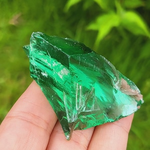 Puede incluir: Un trozo de vidrio verde con una punta afilada, sostenido en una mano. El vidrio es transparente y tiene un ligero tono verde. El fondo está desenfocado y es verde.