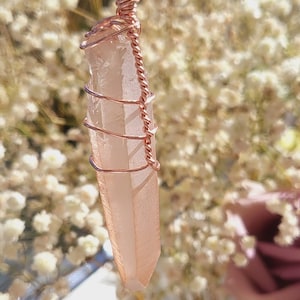 May include: A rose quartz crystal pendant wrapped in copper wire. The pendant is hanging from a black cord.