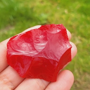 May include: A bright red, irregularly shaped piece of obsidian, a type of volcanic glass, is held in a hand against a green grassy background.