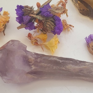 May include: A large, purple amethyst crystal point with a clear, quartz-like tip. The crystal is lying on a white surface with dried flowers scattered around it.