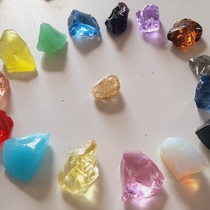 Puede incluir: Una colección de 18 piedras de vidrio de colores dispuestas en círculo sobre una superficie blanca. Las piedras son de varios tonos de rojo, naranja, amarillo, verde, azul, morado y transparente.