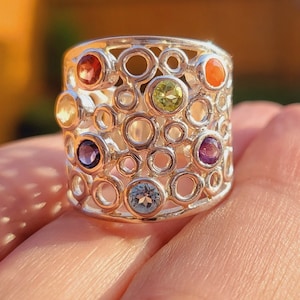 Puede incluir: Un anillo de plata con un diseño circular adornado con piedras preciosas de colores, incluyendo rojo, naranja, amarillo, verde, azul y morado.