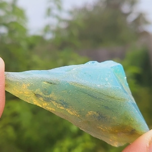 Puede incluir: Un trozo de vidrio de mar con un color iridiscente azul y verde. El vidrio es translúcido y tiene una forma rugosa e irregular.