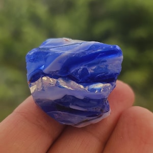May include: A piece of vibrant blue glass with white streaks, held in a hand. The glass has a rough, natural texture and irregular shape. The background is a blurred green, suggesting an outdoor setting.