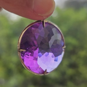 Puede incluir: Un colgante redondo de piedra preciosa púrpura facetada con un engaste de plata.