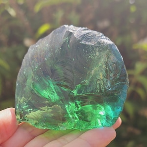 Puede incluir: Un gran trozo de vidrio verde de forma irregular, sostenido en una mano. El vidrio tiene una calidad translúcida, con diferentes tonos de verde y una superficie texturizada. El fondo está borroso con follaje verde.