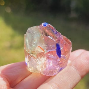 Puede incluir: Un cristal multicolor de forma irregular, con tonos rosas, amarillos y azules. El cristal se sostiene en una mano, con un fondo verde borroso.