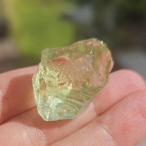 May include: A translucent green crystal with a rough, irregular shape. The crystal is held in a hand, and the background is blurred.
