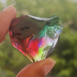 Puede incluir: Una piedra de vidrio en forma de corazón con efecto arcoíris, sostenida entre los dedos. La piedra muestra colores vibrantes como rojo, verde y azul. El fondo es una vista borrosa de follaje verde.
