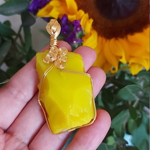 Puede incluir: Un colgante de vidrio amarillo envuelto en alambre dorado, con un diseño de espiral en la parte superior. El colgante se sostiene en una mano, con un girasol y otras flores en el fondo.