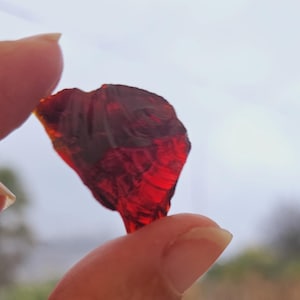 Puede incluir: Un fragmento de piedra preciosa rojo oscuro translúcido, sostenido entre los dedos contra un fondo borroso al aire libre. La gema tiene una apariencia facetada, captando la luz y mostrando su rico color. El tema general son las piedras preciosas naturales.