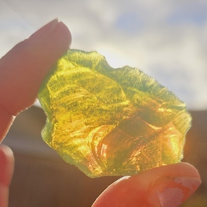 Puede incluir: Un trozo de vidrio translúcido de color amarillo verdoso, de forma irregular, sostenido entre los dedos. La luz del sol brilla a través del vidrio, creando un efecto brillante. El fondo está borroso.
