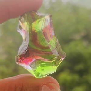 Peut inclure: Un éclat de verre translucide multicolore avec des teintes vertes, roses et jaunes. La forme irrégulière du verre capte la lumière, créant un affichage vibrant de couleurs. L'objet est tenu entre les doigts.