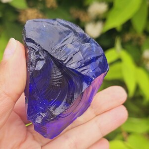 May include: A large, irregularly shaped piece of deep blue glass, held in a hand. The glass has a glossy, reflective surface with internal fractures, and is set against a backdrop of green foliage.