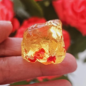 May include: A translucent, amber-colored gemstone with red inclusions is held in a hand. The gemstone has a rough, irregular shape. Red roses are blurred in the background.
