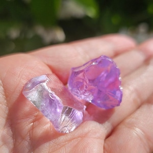May include: Two clear purple crystal chunks in a hand. The crystals are faceted and have a translucent appearance.