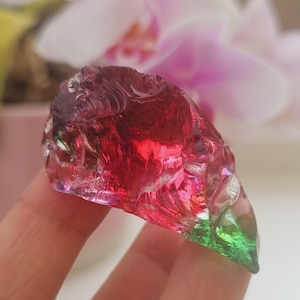 Puede incluir: Un cristal de vidrio multicolor con una textura rugosa y natural. El cristal hace la transición del rojo intenso al verde, con toques de rosa y morado. El cristal se sostiene en una mano, con un fondo floral borroso.