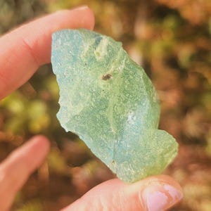 Puede incluir: Una piedra preciosa translúcida de color verde claro sostenida en una mano. La piedra tiene una forma irregular y una textura ligeramente rugosa. El fondo está borroso, con toques de verde y marrón.