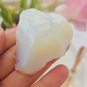 Puede incluir: Una piedra preciosa de ópalo en bruto, de color blanco lechoso y amarillo pálido. La piedra se sostiene en una mano, con flores rosas en el fondo. El ópalo tiene una textura natural y rugosa.
