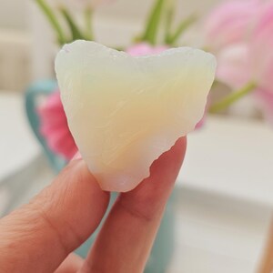 Puede incluir: Se sostiene una piedra translúcida blanquecina de forma irregular. La piedra tiene una textura ligeramente rugosa y un aspecto suave y brillante. Hay tulipanes rosas en el fondo.