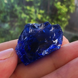 May include: A raw, deep cobalt blue glass chunk with an irregular, jagged shape and a translucent, reflective surface. The piece features sharp, fractured edges and a smooth, glossy texture that catches light, displaying varying shades of blue.