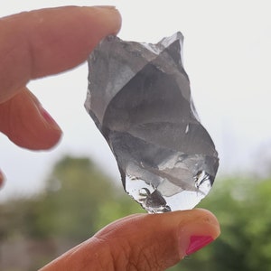 Puede incluir: Una roca transparente, facetada, similar a un cristal, con una forma irregular y rugosa. La roca está siendo sostenida por un dedo con esmalte de uñas rosa.