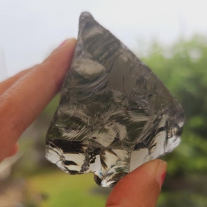 Könnte beinhalten: Ein klares, facettierter, dreieckiger Obsidianstück mit einer grauen, wolkigen Optik. Der Obsidian wird in einer Hand gehalten.