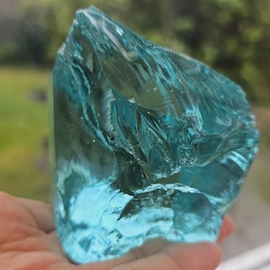 May include: A large, clear, teal-coloured glass shard with many facets. The shard is being held in a hand.
