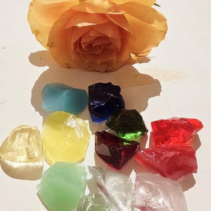 Peut inclure: Une composition de morceaux de verre colorés de formes irrégulières, bleus, jaunes, verts, rouges et transparents, avec une rose pêche en arrière-plan. Les morceaux de verre sont disposés sur une surface blanche.